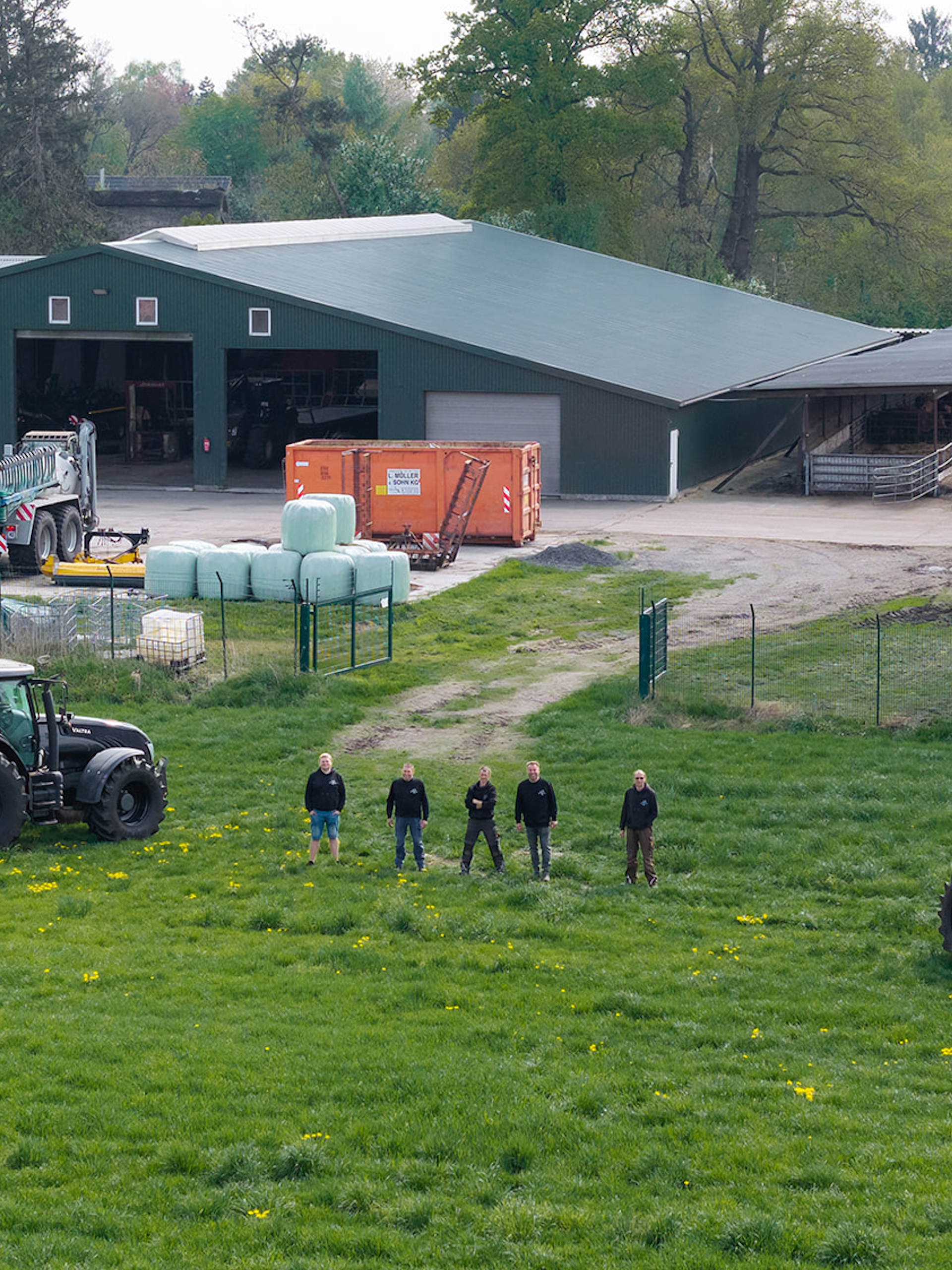 Auf dem Bild sieht man eine Halle für Landwirtschaftsmaschinen aus einer erhöhten Position abgelichtet, sowie fünf Personen die auf einer Weide stehen.